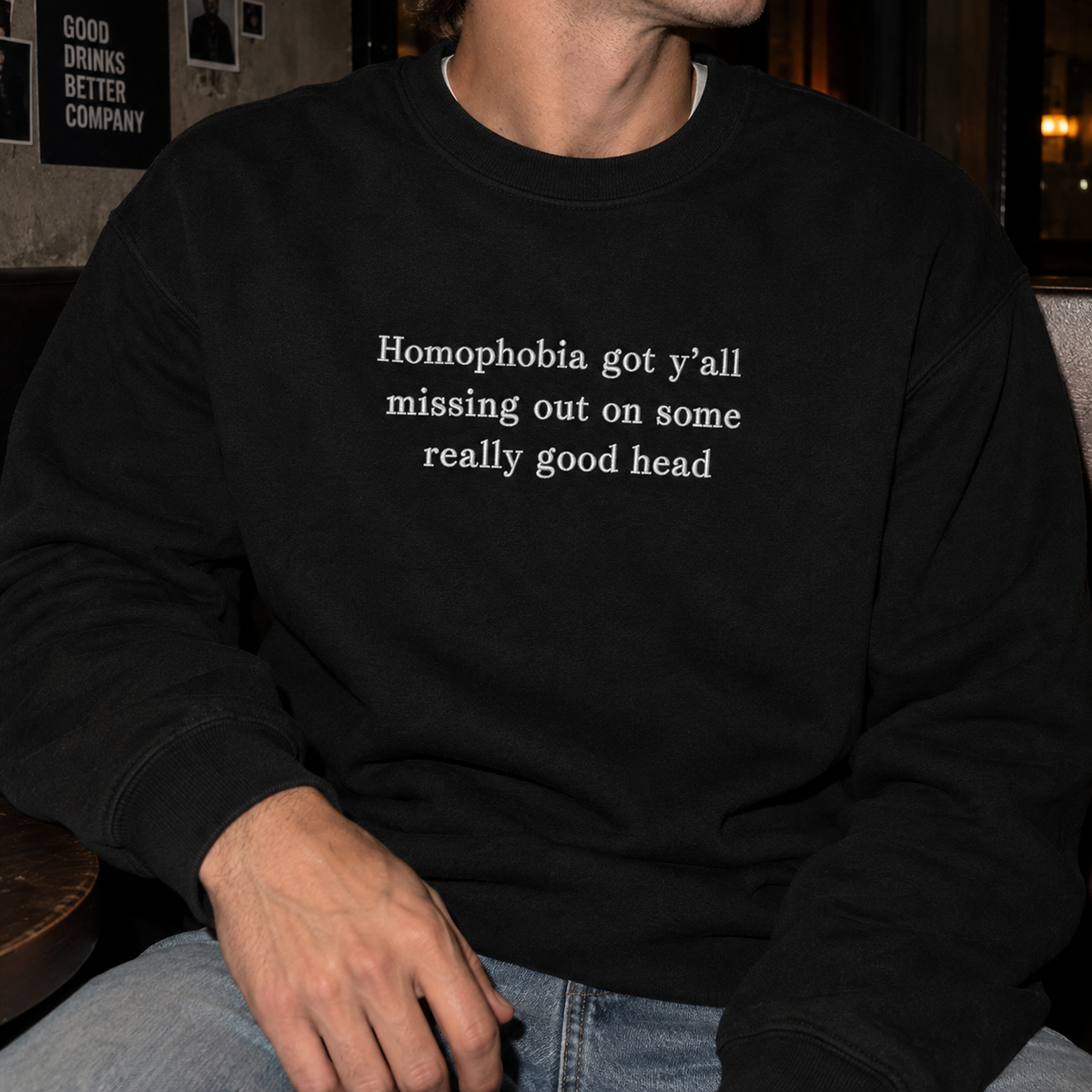 A person wearing a black crewneck sweatshirt with the white printed text 'Homophobia got y'all missing out on some really good head', paired with blue jeans in a dimly lit indoor setting.