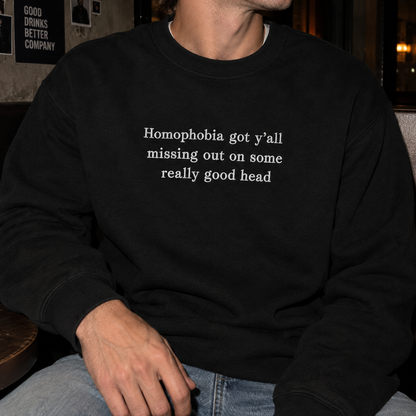 A person wearing a black crewneck sweatshirt with the white printed text 'Homophobia got y'all missing out on some really good head', paired with blue jeans in a dimly lit indoor setting.