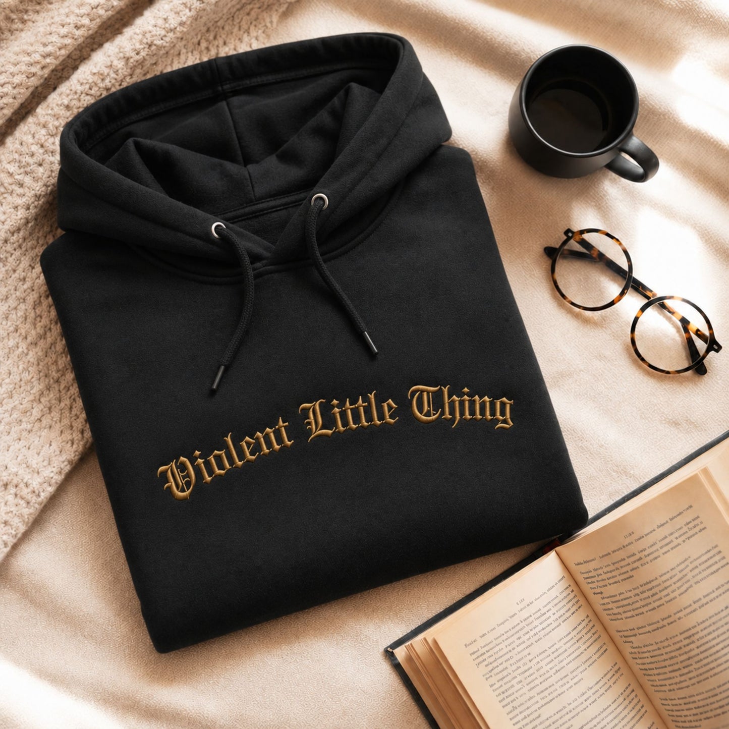 A folded black hoodie with the phrase 'Violent Little Thing' embroidered in gold across the chest, styled on a beige blanket with a mug, glasses, and an open book nearby.