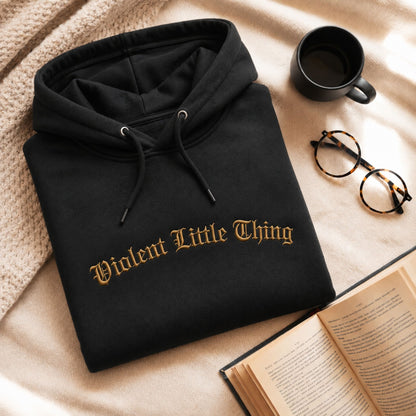 A folded black hoodie with the phrase 'Violent Little Thing' embroidered in gold across the chest, styled on a beige blanket with a mug, glasses, and an open book nearby.