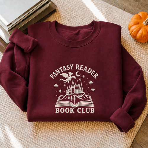 A folded maroon sweatshirt with a white graphic reading 'FANTASY READER BOOK CLUB' and showing an open book, castle, dragon, and stars, styled on a woven beige surface with books and a small pumpkin nearby.