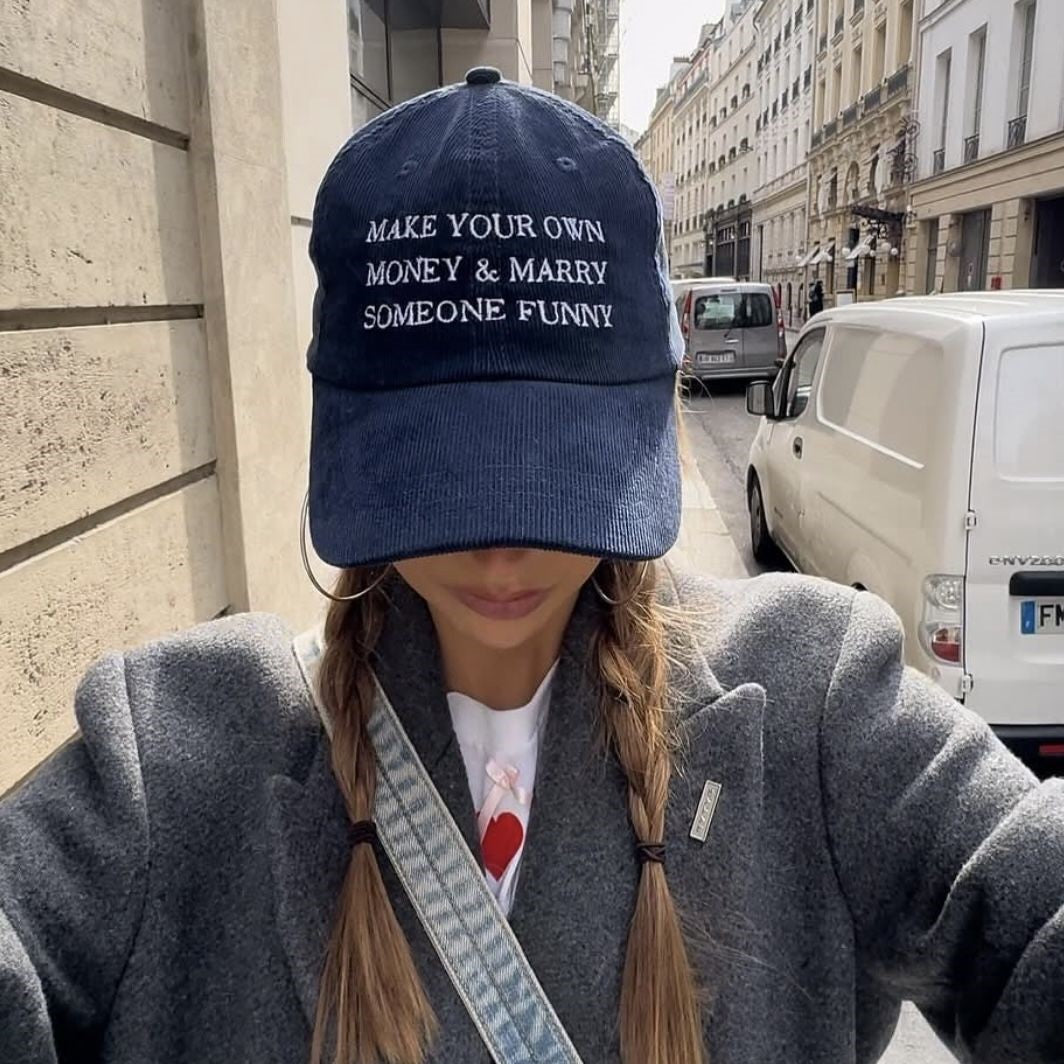 A person taking a selfie on a city street wearing a navy blue baseball cap with white embroidered text that reads 'MAKE YOUR OWN MONEY & MARRY SOMEONE FUNNY'.