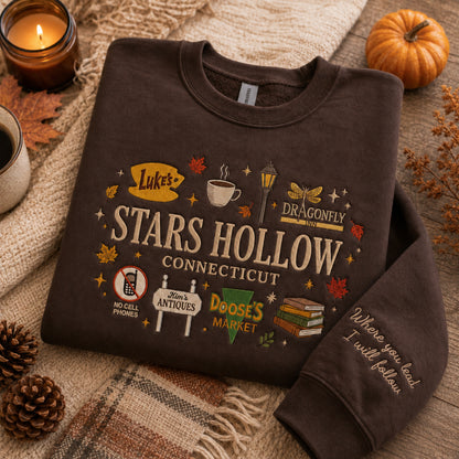 A dark brown crewneck sweatshirt featuring a colorful graphic print for 'Star's Hollow Connecticut' with pop culture references, styled in a cozy flat-lay setting with autumn props like a candle, pumpkin, and plaid blanket.