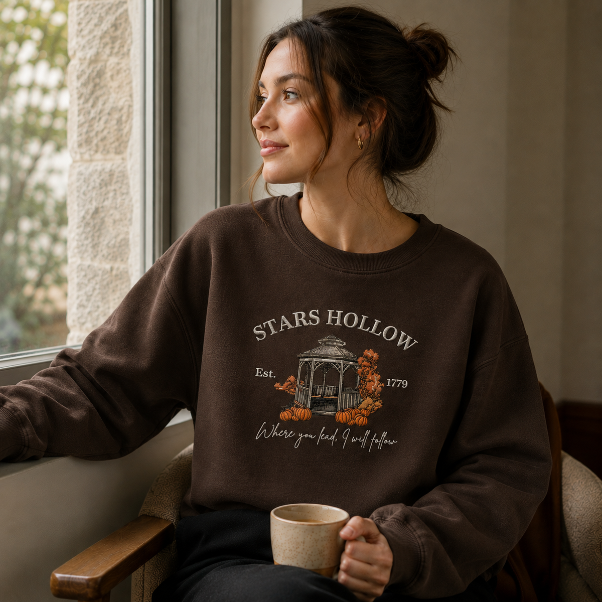 A woman sitting in a chair by a window wearing a dark brown crewneck sweatshirt with a 'STARS HOLLOW' graphic. She is holding a ceramic mug.