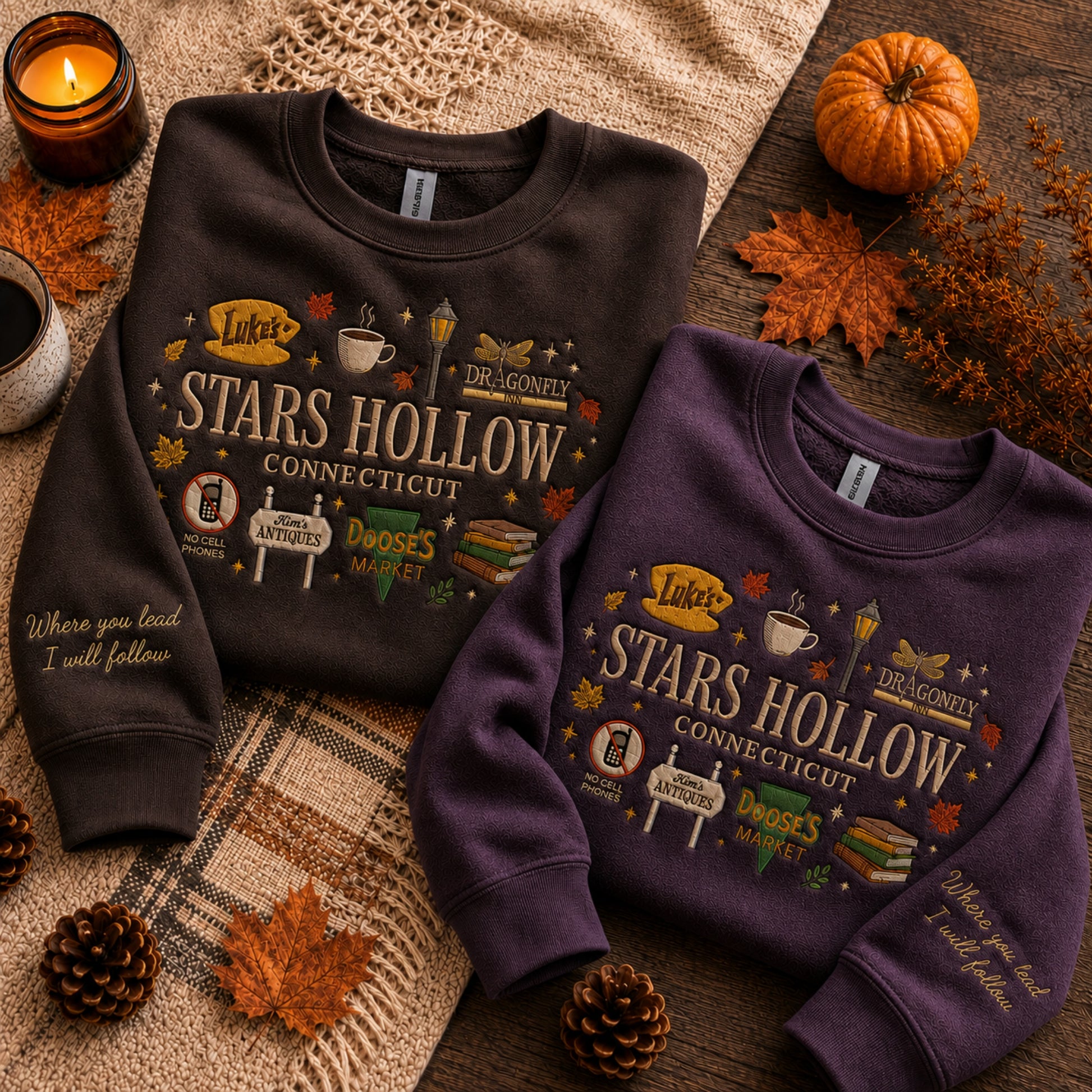 Two crewneck sweatshirts laid flat on a rustic autumn-themed surface. One sweatshirt is dark charcoal brown and the other is deep purple, both featuring a large 'Luka's Stars Hollow Connecticut' graphic with book, coffee, and fall leaf motifs.