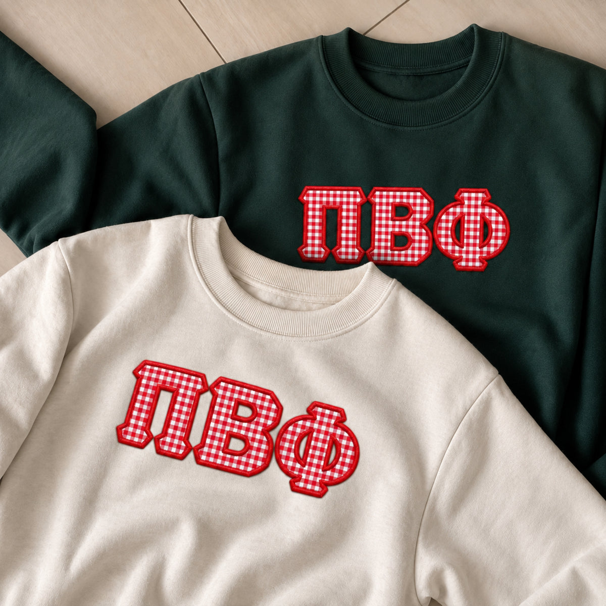 Two crewneck sweatshirts laid flat on a light wood floor: one cream and one dark forest green, both featuring large red-and-white gingham block letters outlined in red across the chest.