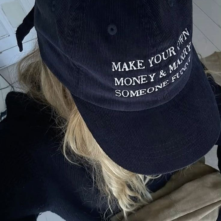 A person wearing a black corduroy baseball cap with white embroidered text that reads 'MAKE YOUR OWN MONEY & MARRY SOMEONE FURTHER'.