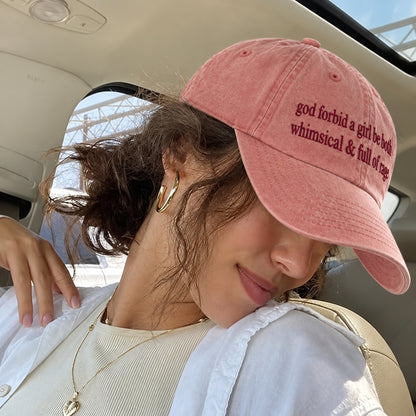 A woman in a car wearing a dusty pink baseball cap with embroidered text that reads 'god forbid a girl be born whimsical & full of care'.