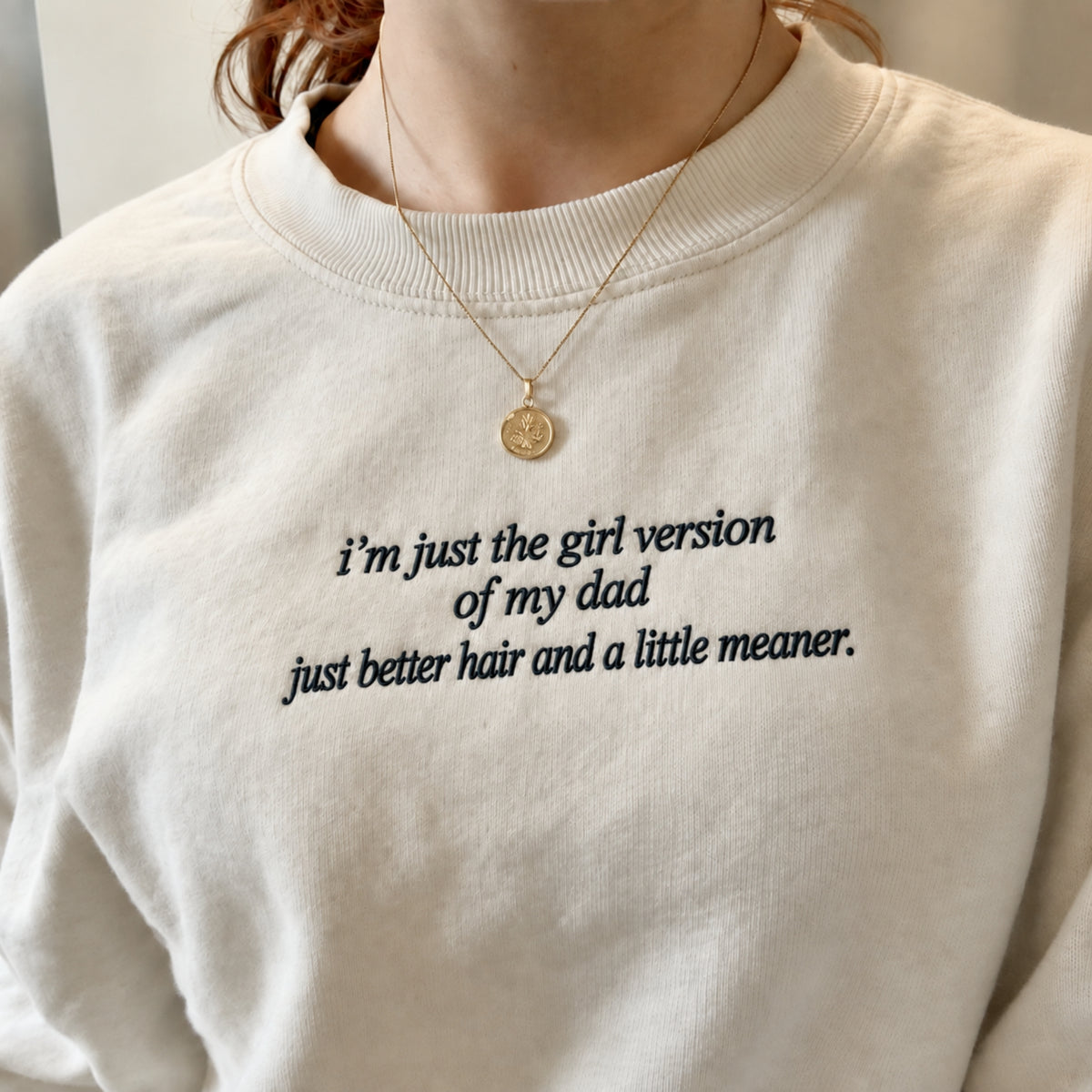A close-up of a person wearing a cream-colored crewneck sweatshirt with black embroidered text that reads 'i'm just the girl version of my dad just better hair and a little meaner.' A delicate gold necklace with a small round pendant is also visible.