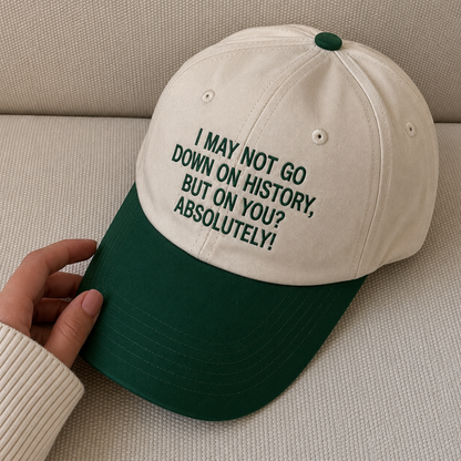 A baseball cap with an off-white crown and a dark green brim, held by a hand. The front of the cap features dark green embroidered text that reads, 'I MAY NOT GO DOWN ON HISTORY, BUT ON YOU? ABSOLUTELY!'.