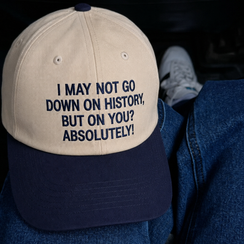 A beige and navy blue baseball cap with the text 'I MAY NOT GO DOWN ON HISTORY, BUT ON YOU? ABSOLUTELY!' embroidered in navy on the front, shown resting on blue denim next to a white sneaker.