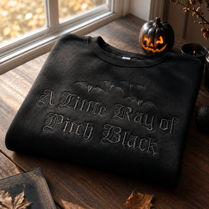 Black gothic crewneck sweatshirt with detailed bat embroidery and A Little Ray of Pitch Black lettering, folded on a wooden table with Halloween-inspired decorations and warm natural lighting.