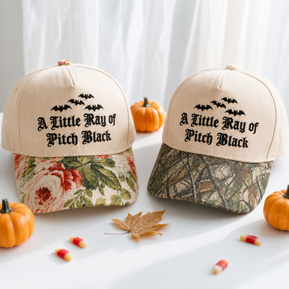 Two beige baseball caps with black gothic text reading 'A Little Ray of Pitch Black' and bat silhouettes, styled on a white surface with small pumpkins, candy corn, and a leaf. The left cap has a floral brim, and the right cap has a camouflage brim.