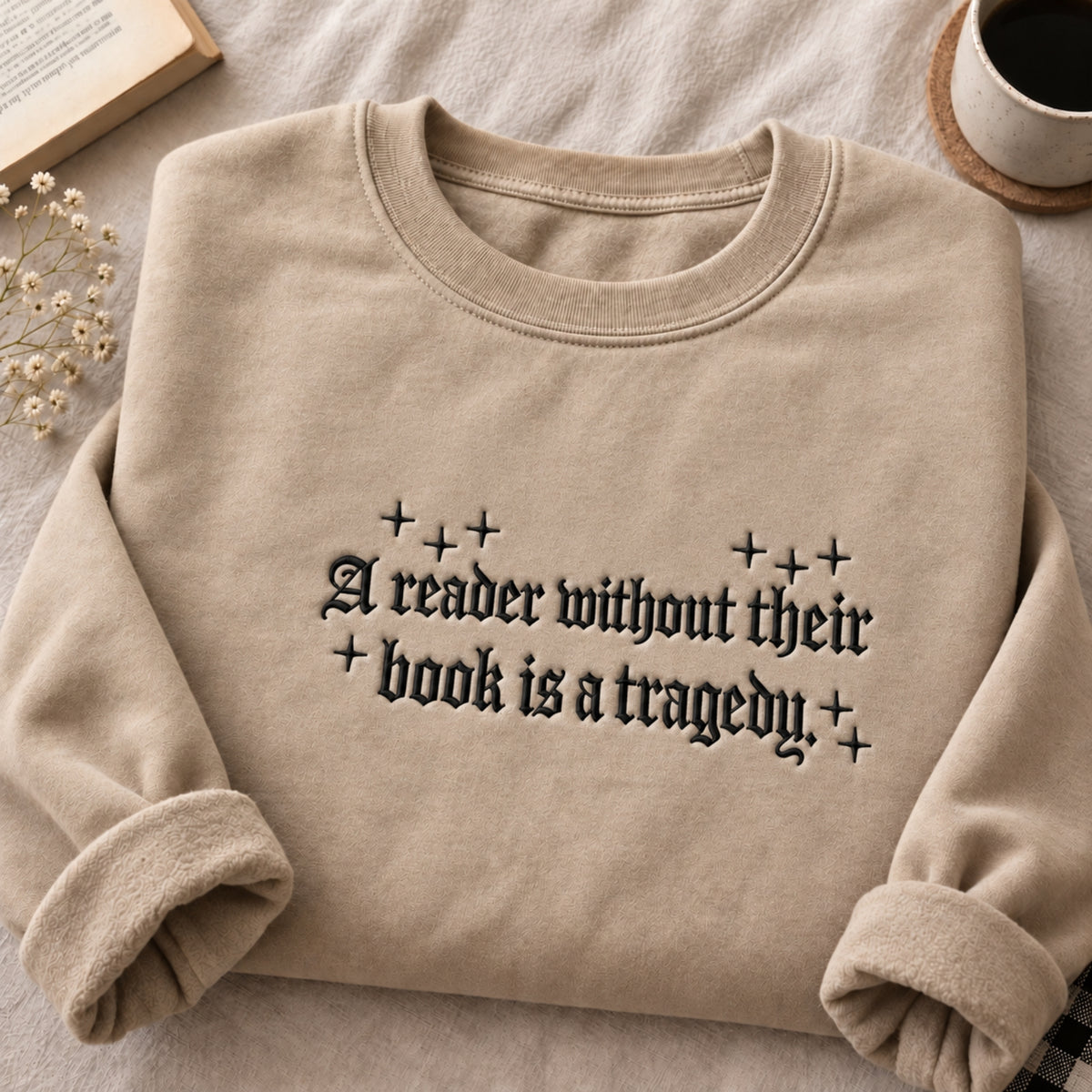 A beige crewneck sweatshirt with rolled cuffs, featuring the text 'A reader without their book is a tragedy' in a black gothic font with decorative plus signs. The sweatshirt is styled in a cozy flat-lay setting on a neutral fabric background with a book, dried flowers, and a coffee cup.