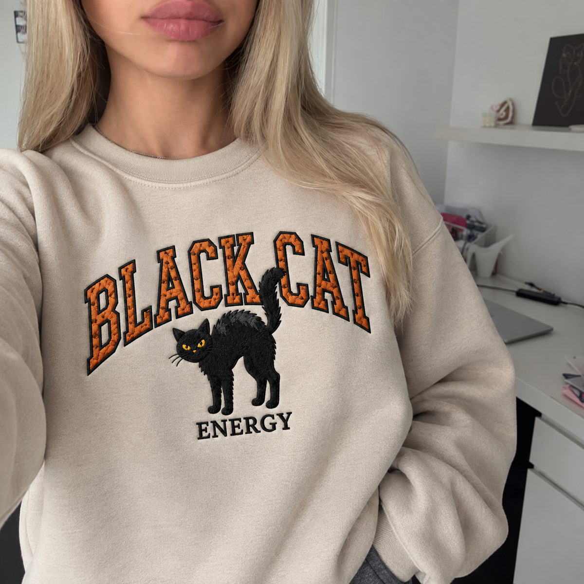 A woman wearing a cream-colored sweatshirt with a graphic that reads 'BLACK CAT ENERGY' featuring an embroidered black cat. The photo is taken in a casual indoor setting.