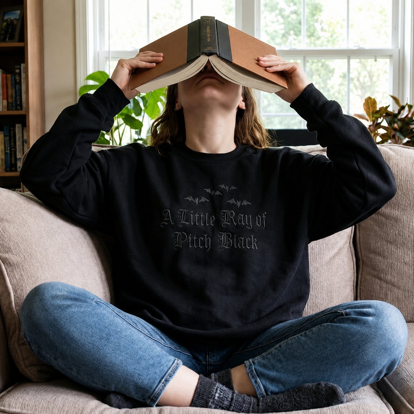 Person relaxing on a couch while wearing an oversized goth aesthetic crewneck embroidered with the A Little Ray of Pitch Black design and subtle black bat embroidery.
