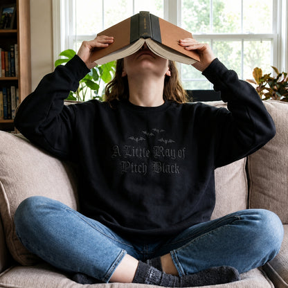 Person relaxing on a couch while wearing an oversized goth aesthetic crewneck embroidered with the A Little Ray of Pitch Black design and subtle black bat embroidery.