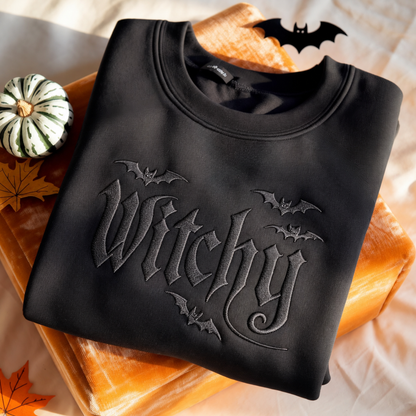A folded black crewneck sweatshirt with a tonal gothic 'Witchy' text design and bat motifs, styled on a stack of orange books with autumn leaves and a small pumpkin.