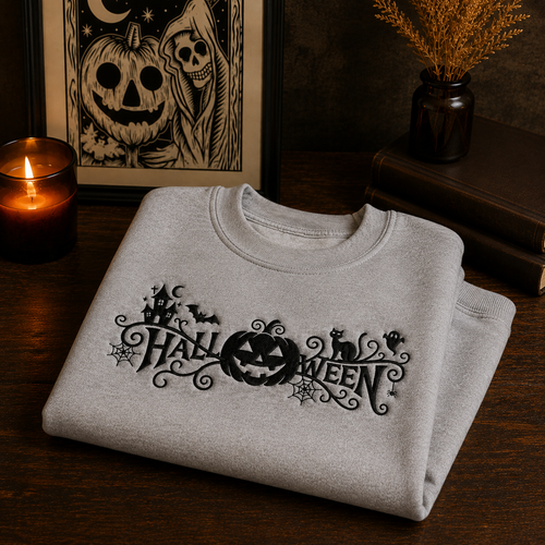 A folded light gray crewneck sweatshirt with a black embroidered 'Halloween' graphic sits on a dark wooden table. The background is styled with a burning candle, a framed skeleton and pumpkin print, and a small vase of dried grass.