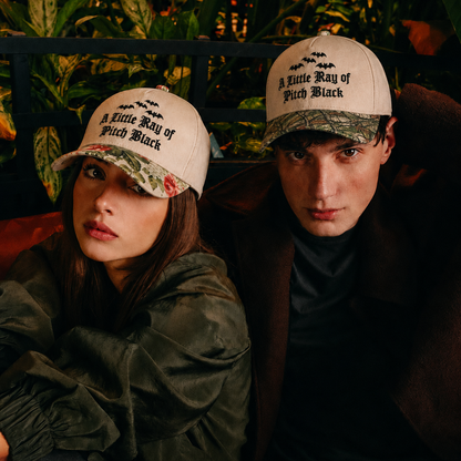 Two models wearing matching beige baseball caps with black text reading 'A Little Way of Pitch Black' and camouflage brims, posed in a dark, plant-filled setting.