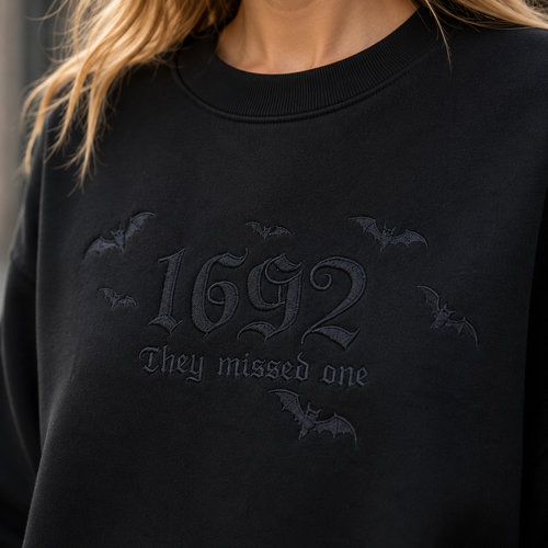 Close-up of a person wearing a black crewneck sweatshirt with tonal dark gray gothic text reading '1692 They missed one' and small bat silhouettes, shown against a softly blurred outdoor background.