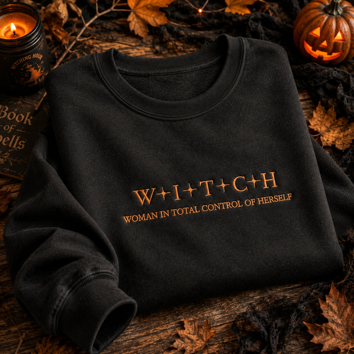 A black crewneck sweatshirt with gold foil text reading 'W+I+T+C+H' and 'WOMAN IN TOTAL CONTROL OF HERSELF' laid flat on a rustic wooden surface surrounded by Halloween props including a candle, spiderweb, leaves, and a jack-o'-lantern.