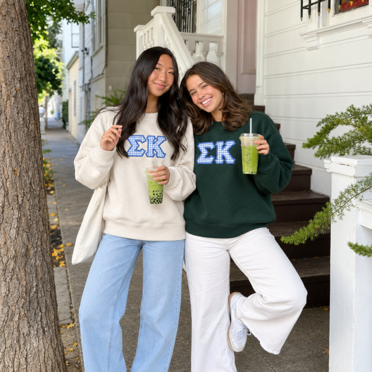 Two women standing on a front porch outdoors, each wearing a sweatshirt with a blue Greek letter 'ΣΚ' graphic. The woman on the left wears a cream sweatshirt with light blue jeans and holds a green boba drink.