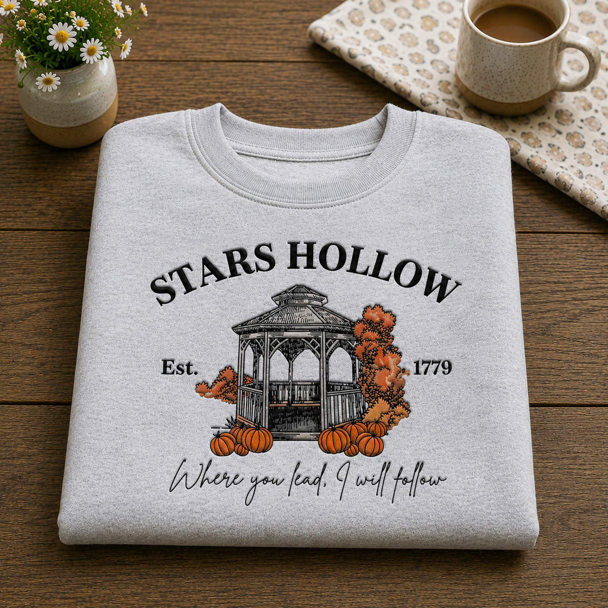 A light heather gray folded T-shirt with a fall-themed graphic reading 'STARS HOLLOW' and 'Where you lead, I will follow,' displayed on a wooden surface with a small flower pot, mug, and patterned cloth nearby.