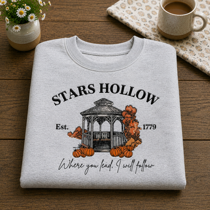 A light heather gray folded T-shirt with a fall-themed graphic reading 'STARS HOLLOW' and 'Where you lead, I will follow,' displayed on a wooden surface with a small flower pot, mug, and patterned cloth nearby.
