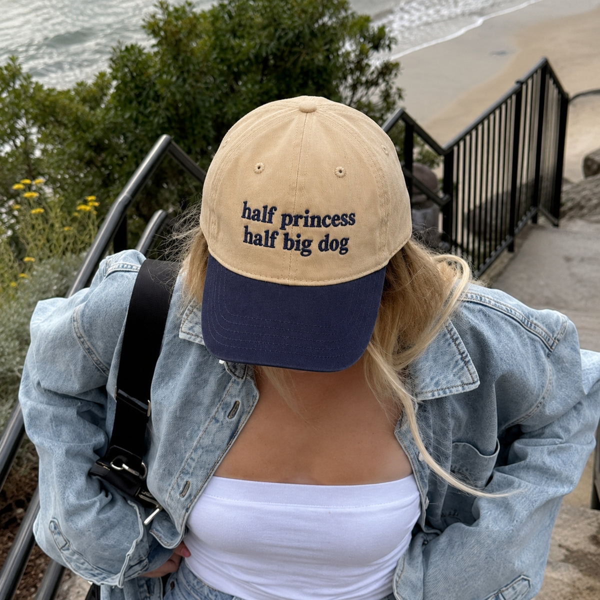 A person outdoors near a beach and railing wears a beige and navy baseball cap with the embroidered text 'half princess half big dog'.