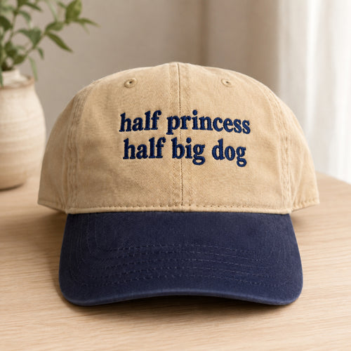 A two-tone baseball cap with a beige crown and a navy blue brim, featuring the embroidered text 'half princess half big dog' in navy blue on the front. The cap is displayed on a light wood surface with a blurred potted plant in the background.