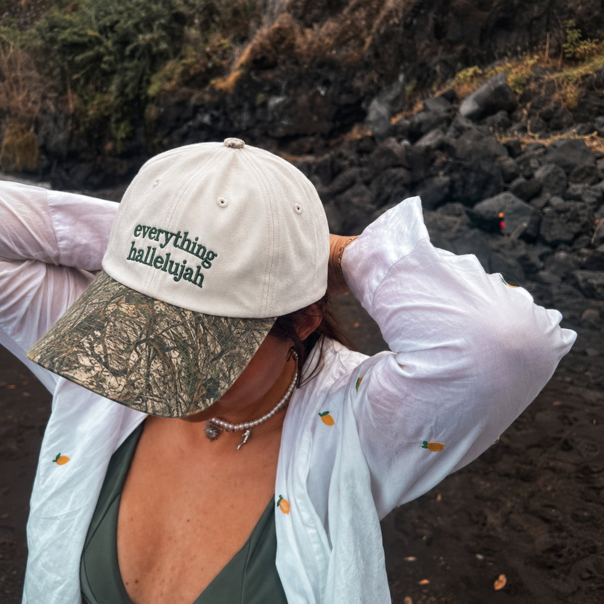 A person outdoors on a rocky beach wearing a cream baseball cap with 'everything hallelujah' embroidered on the front and a camouflage visor, along with a white long-sleeve shirt and dark green top.