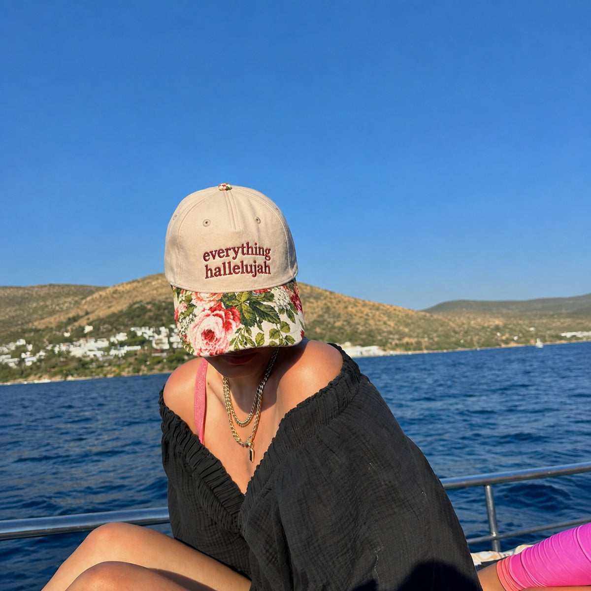 A person sitting outdoors on a boat or dock wearing a beige baseball cap with the words 'everything hallelujah' in red embroidery and a floral brim, with blue water and hills in the background.