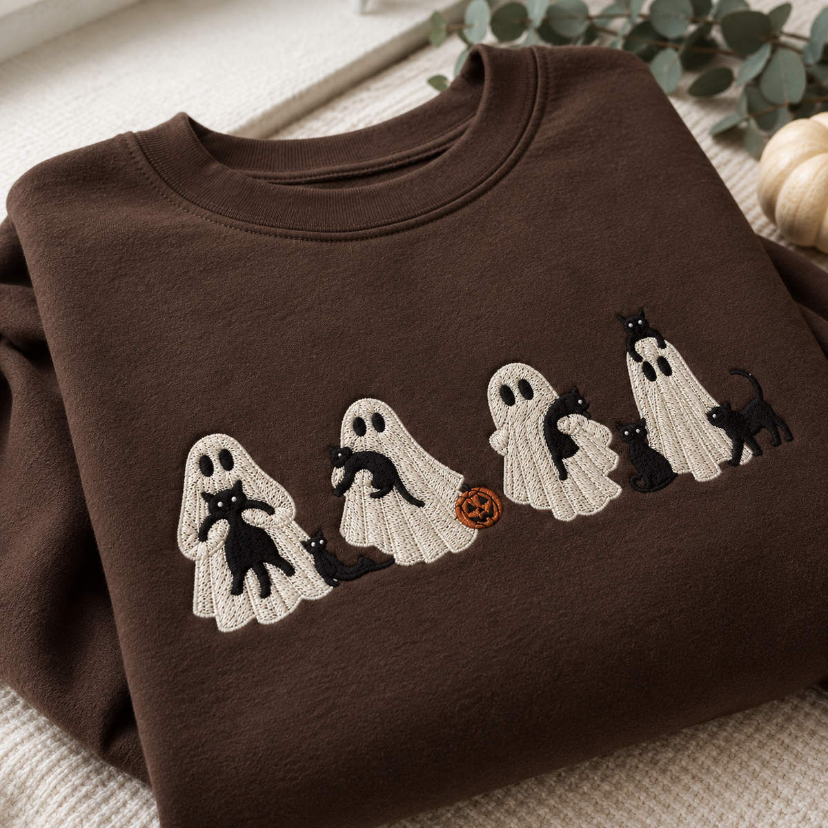 A folded brown crewneck sweatshirt featuring a knitted-style embroidered graphic of four white ghosts carrying black cats, with one ghost holding an orange pumpkin, displayed on a neutral textured surface.