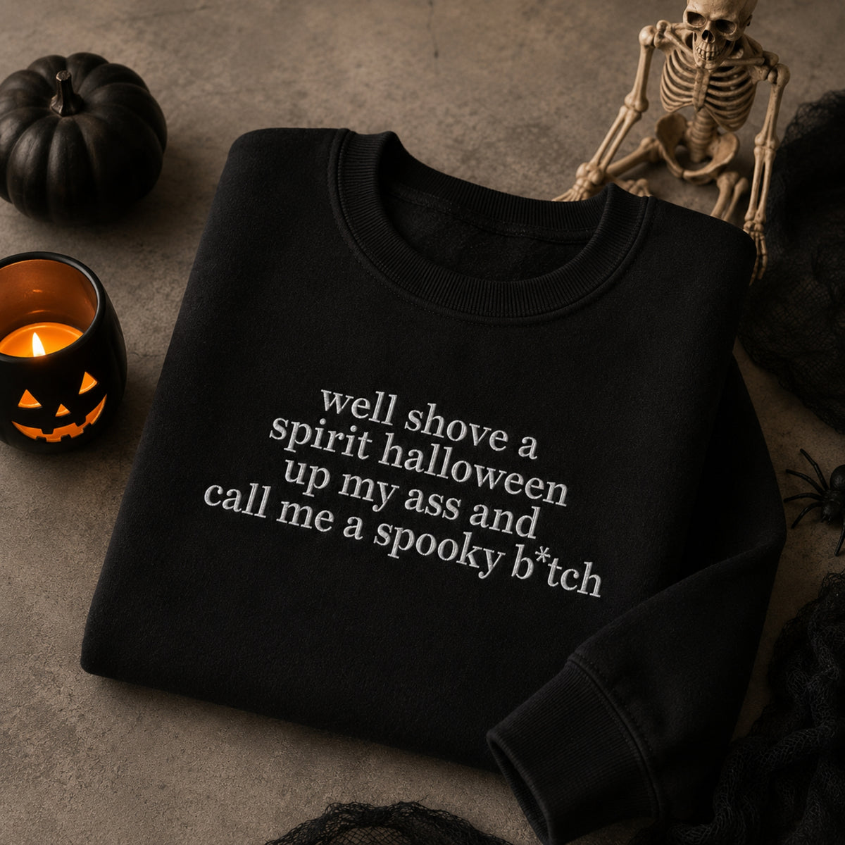 A black crewneck sweatshirt with white text reading 'well shove a spirit halloween up my ass and call me a spooky b*tch' is displayed in a Halloween-themed flat lay with a pumpkin, skeleton, and candle.
