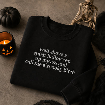 A black crewneck sweatshirt with white text reading 'well shove a spirit halloween up my ass and call me a spooky b*tch' is displayed in a Halloween-themed flat lay with a pumpkin, skeleton, and candle.