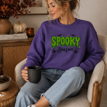 A woman sitting in a cozy armchair wearing a purple sweatshirt with a green and black 'Spooky Season' graphic, paired with light blue jeans and holding a black mug.