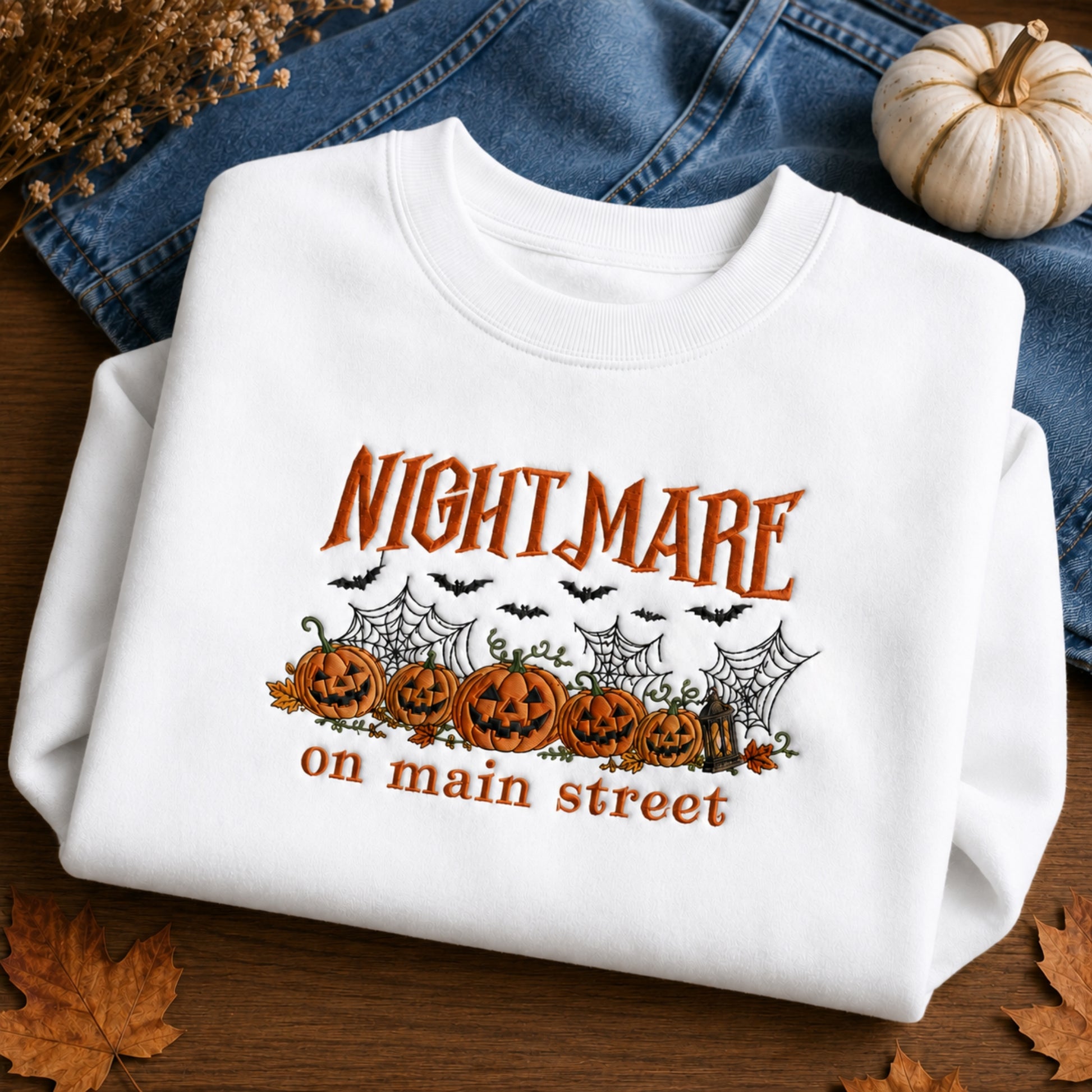 A folded white crewneck sweatshirt with a Halloween graphic reading 'NIGHTMARE on main street' above jack-o'-lanterns, cobwebs, bats, and leaves, styled on a wooden surface with blue jeans, a small pumpkin, and dried leaves.