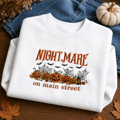 A folded white crewneck sweatshirt with a Halloween graphic reading 'NIGHTMARE on main street' above jack-o'-lanterns, cobwebs, bats, and leaves, styled on a wooden surface with blue jeans, a small pumpkin, and dried leaves.