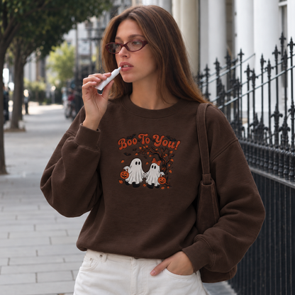 A woman wearing a brown crewneck sweatshirt with a 'Boo To You!' Halloween graphic featuring two ghosts and pumpkins, standing on a city sidewalk.