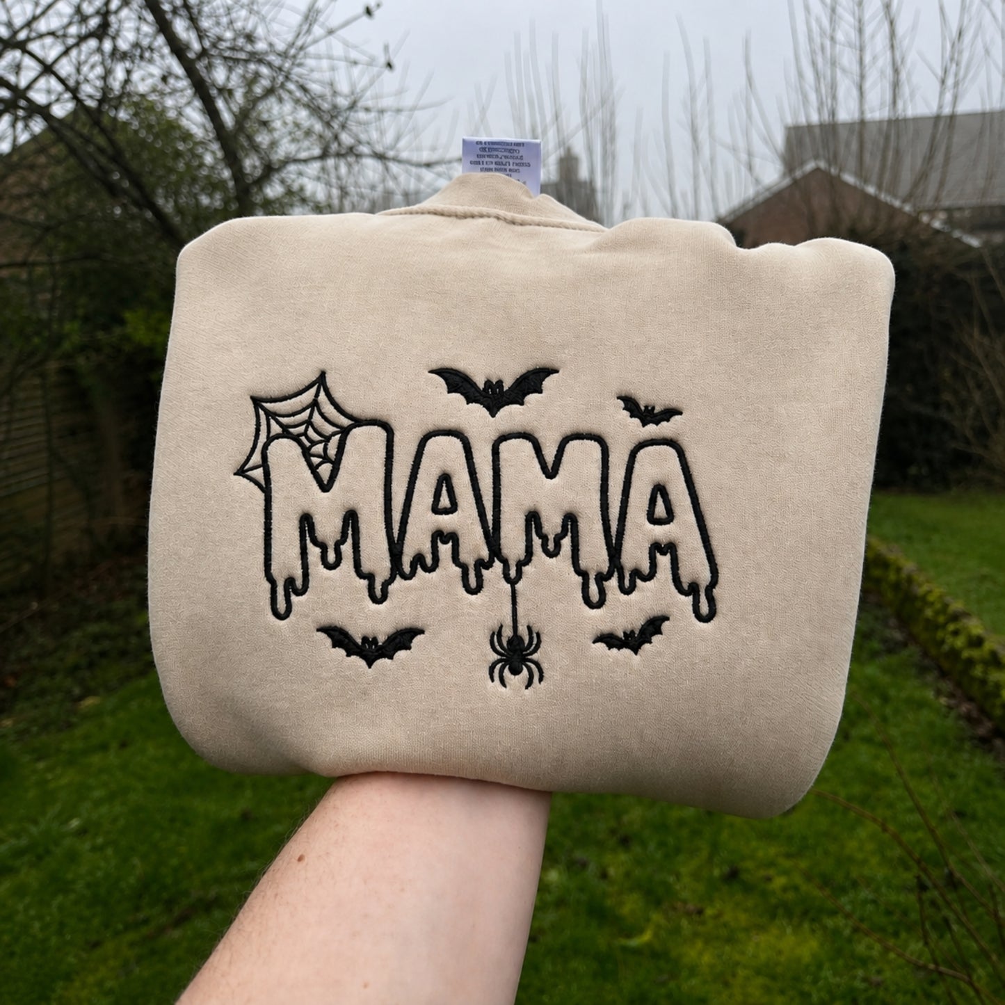 A beige fleece sweatshirt cover held up by a hand outdoors. The cover features black embroidered text reading 'MAMA' in a dripping font, decorated with black spiderwebs and bats.