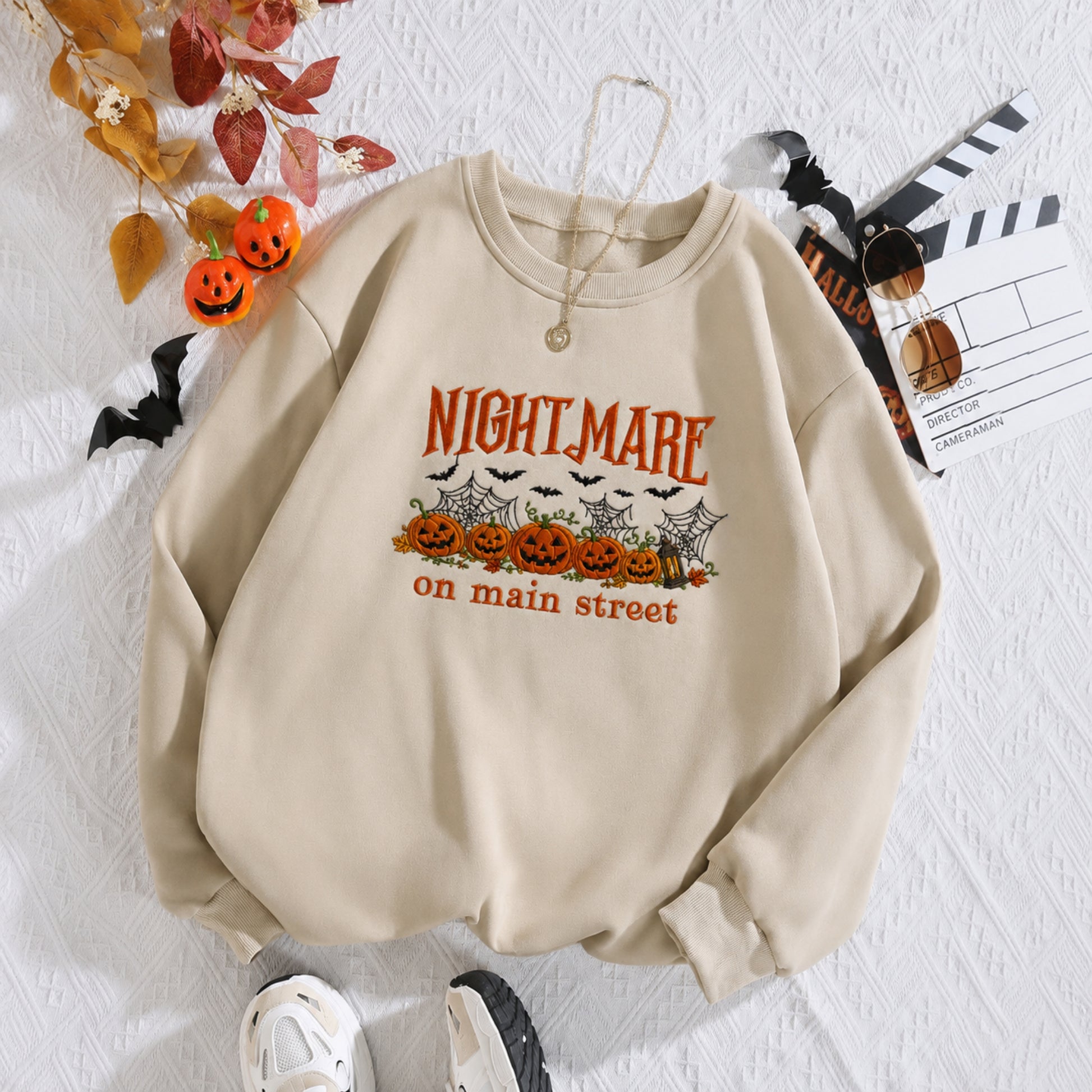 A beige crewneck sweatshirt with a Halloween graphic print reading 'NIGHTMARE on main street' above jack-o'-lanterns, bats, and spiderwebs, styled in a flat lay with autumn leaves, a clapperboard, and sneakers on a white textured background.