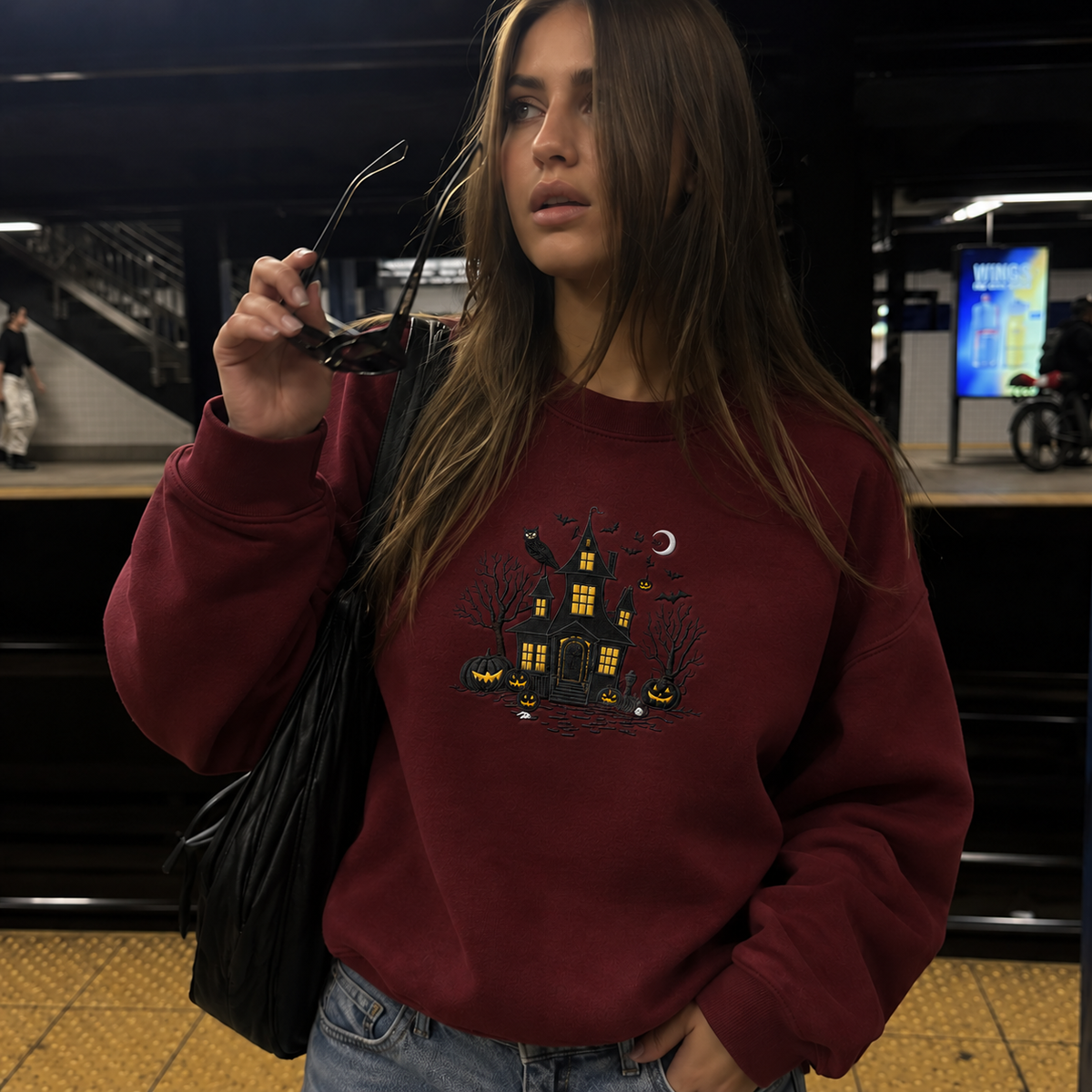 A woman wearing a burgundy crewneck sweatshirt with a black and yellow haunted house graphic, standing in a subway station and holding sunglasses.