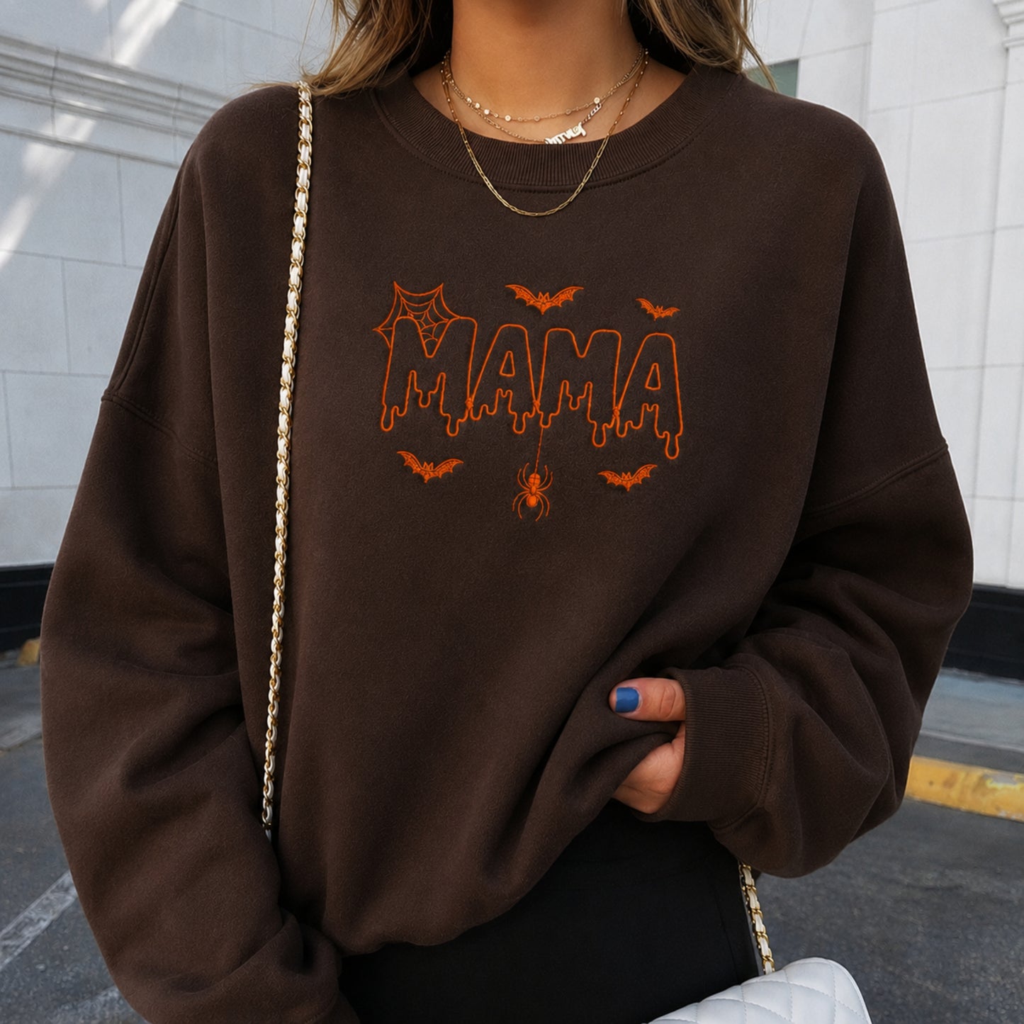 A woman wearing a brown crewneck sweatshirt with an orange 'MAMA' graphic and spider-themed details, styled with gold necklaces and a quilted bag.