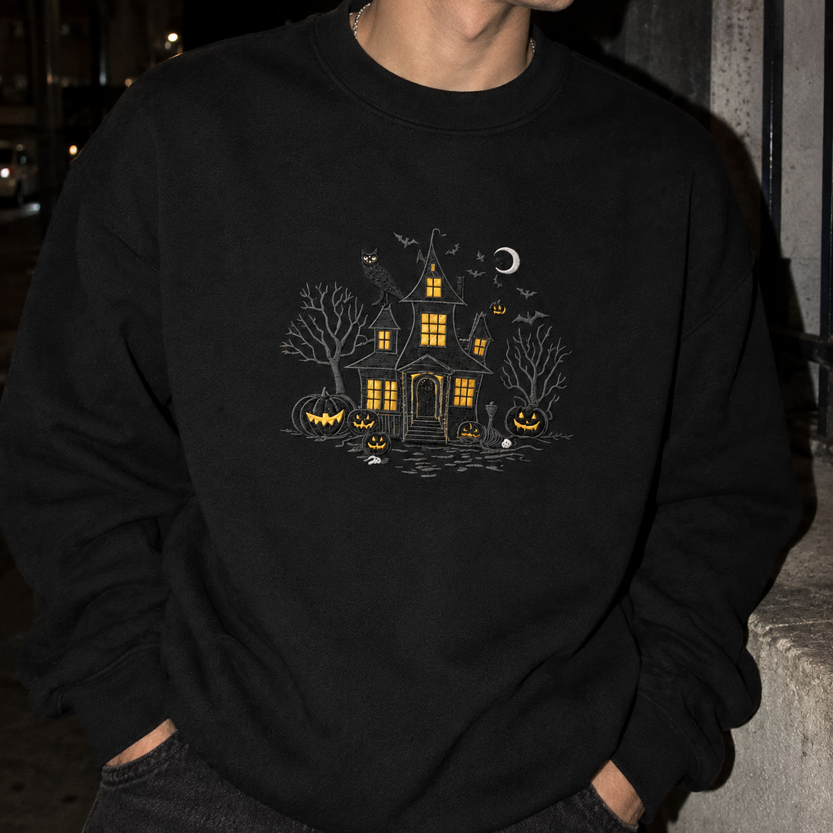 A person wearing a black crewneck sweatshirt with a detailed Halloween graphic on the chest, featuring a haunted house, jack-o'-lanterns, bats, and a crescent moon in yellow, white, and gray.