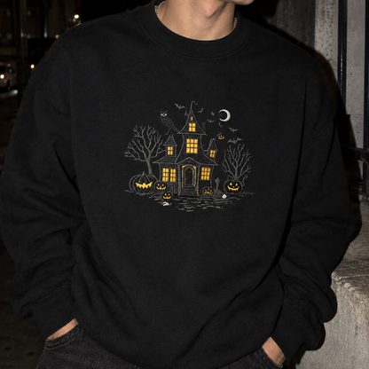 A person wearing a black crewneck sweatshirt with a detailed Halloween graphic on the chest, featuring a haunted house, jack-o'-lanterns, bats, and a crescent moon in yellow, white, and gray.