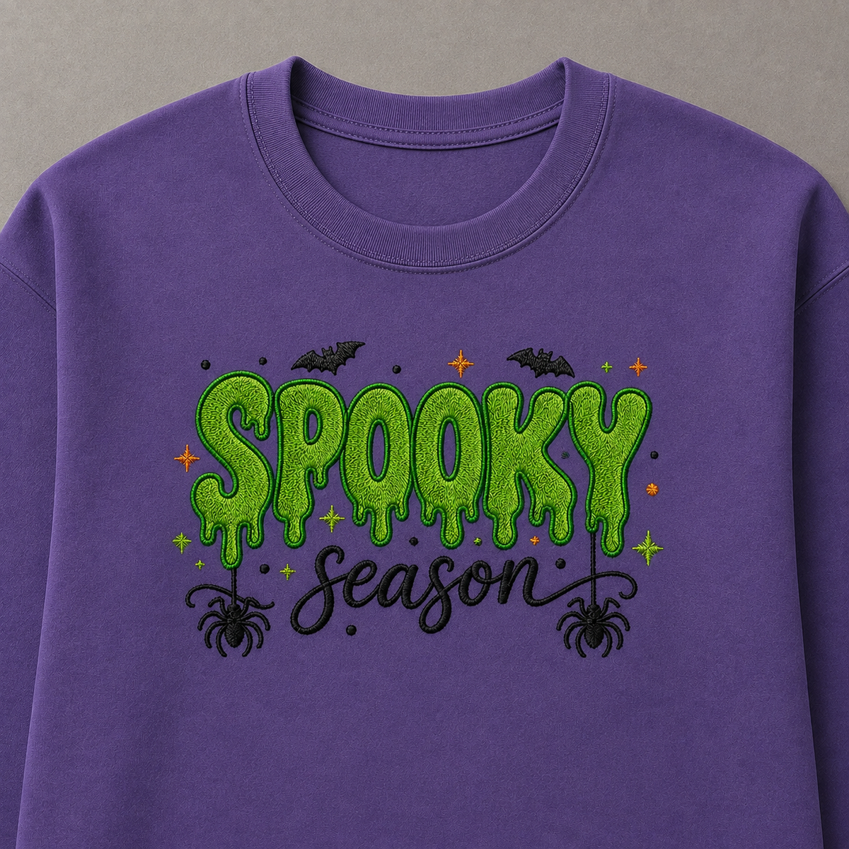 A close-up of a purple crewneck sweatshirt featuring a 'Spooky Season' graphic on the front. The graphic includes green glittery dripping letters, black bats, spiders, and stars, set against a neutral gray background.