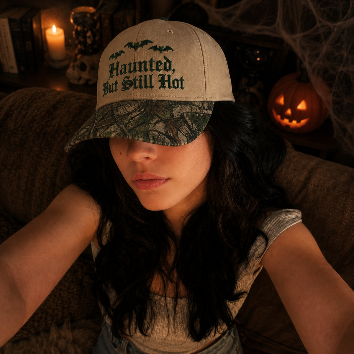 A woman wearing a beige baseball cap with 'Haunted, But Still Hot' and bat graphics on the front panel, paired with a camouflage visor, in a dimly lit Halloween-themed room.