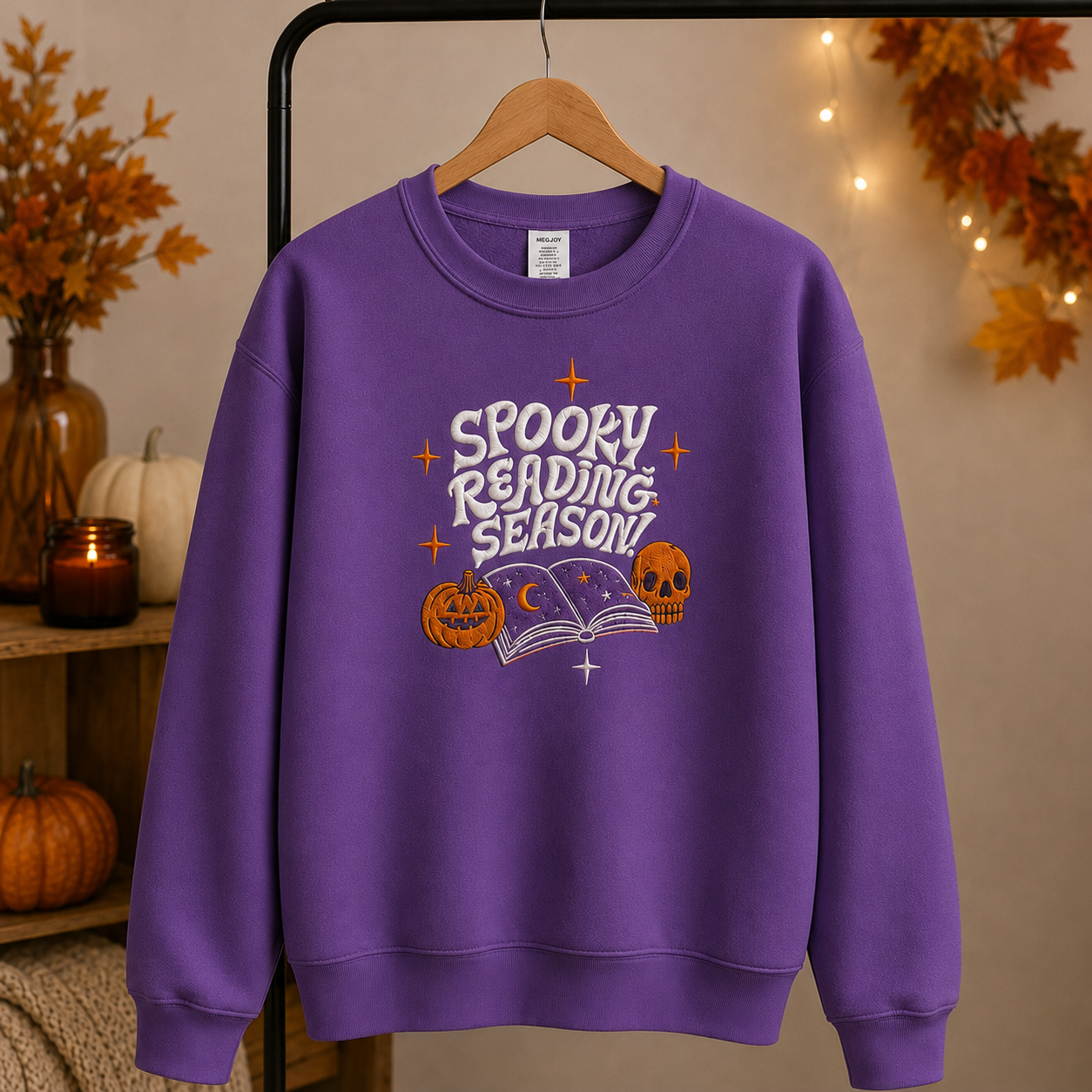 A purple crewneck sweatshirt hanging on a wooden hanger, featuring a 'Spooky Reading Season!' graphic with pumpkins, a book, and stars. The sweatshirt is displayed on a clothing rack against a warm, cozy background with autumn decorations, string lights, and pumpkins.