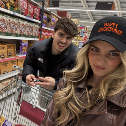 A man and a woman in a grocery store aisle. The woman in the foreground wears a black baseball cap with orange text reading 'HAPPY COCKTOBER' and a brown leather jacket, while the man behind her wears a black puffer jacket.